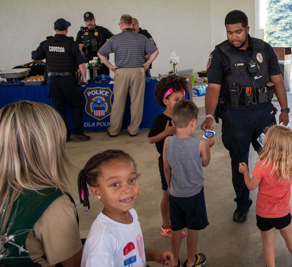 A dark skinned man in a police uniform hands out stickers to several children in a pavilion.