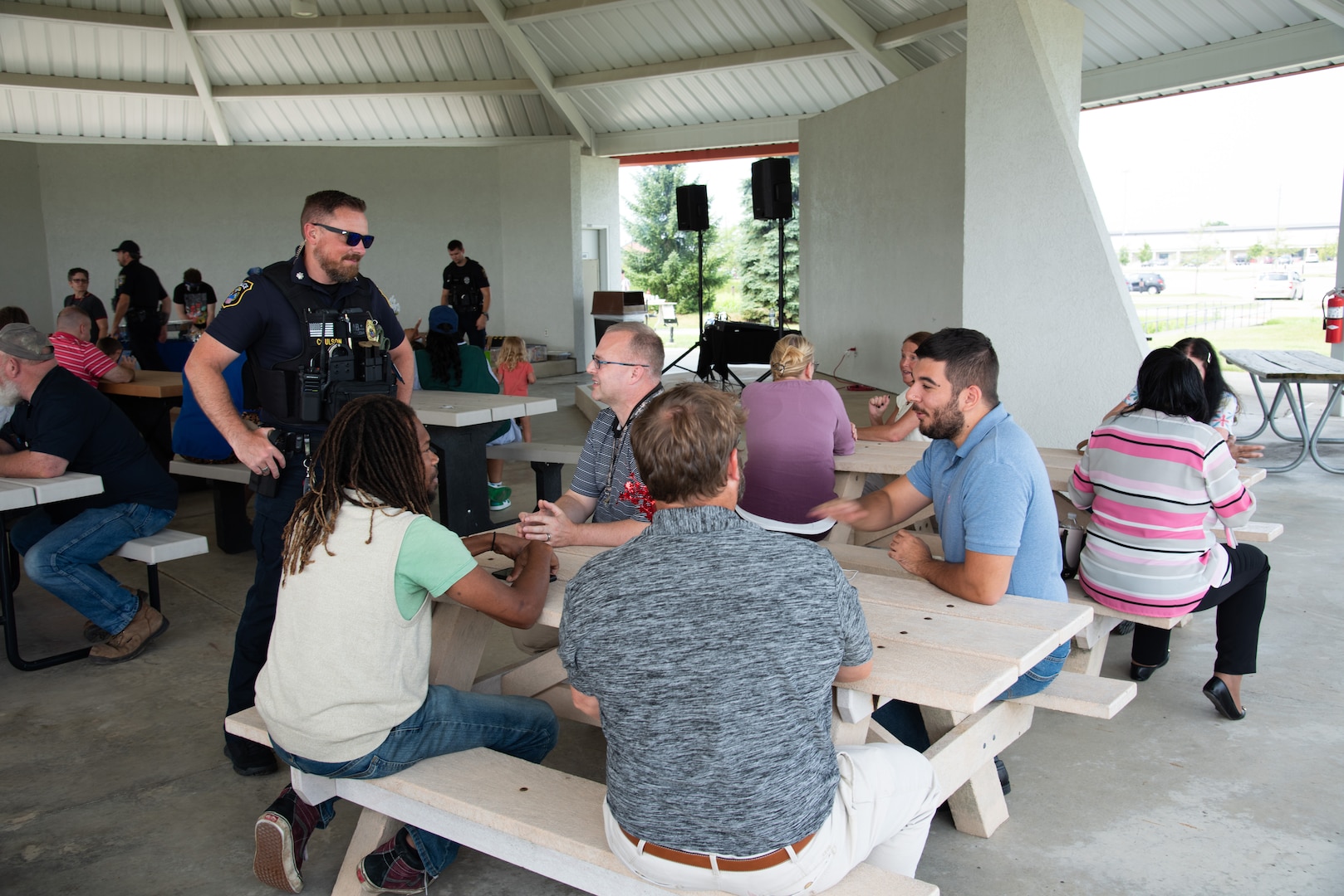 A light skinned man in a police uniform with sunglasses on chats with several people at a picnic table in a pavilion.