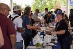 A light skinned woman wearing a cardboard police hat and a black T-shirt serves food to a group of people in a picnic pavilion.