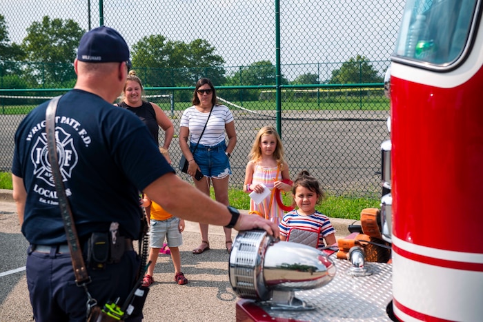 A firefighter in a blue shirt rests his hand on one of the lights of a firetruck as to explain to the children standing in front of him what it is and how it works.