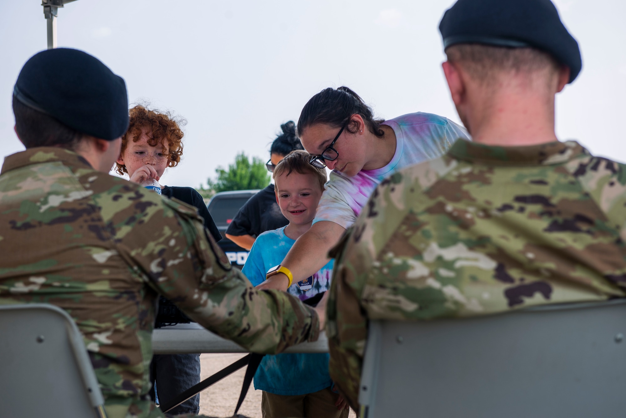Two security forces airmen sit at a table with 2 children and their mother as the mother points to something on the table that makes one of the children smile.