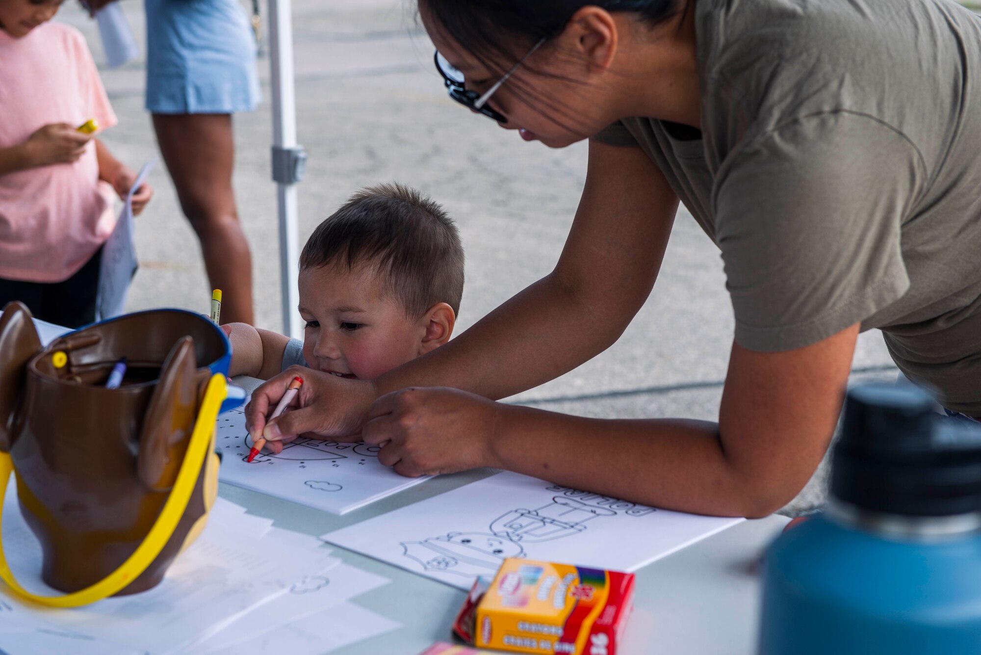 A young child with his head barely over the plastic table, colors a coloring sheet next to his mom on the right who is wearing sunglasses and an ocp shirt.