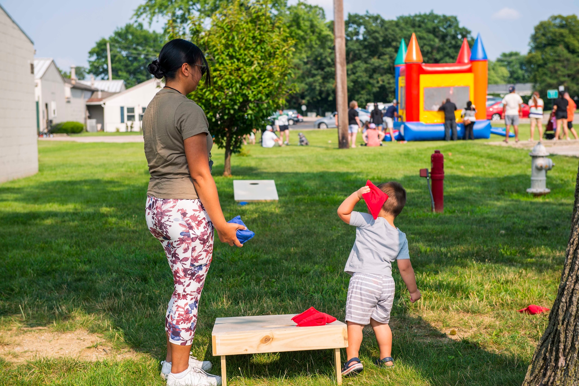 A woman with an ocp shirt and colorful pants stands over a cornhole board holding a blue bag while her very young son stands on the other side of the board and throws a red bag at the opposing board underneath a tree in a grassy lot with a bouncy house in the background that has many children and parents standing around it.