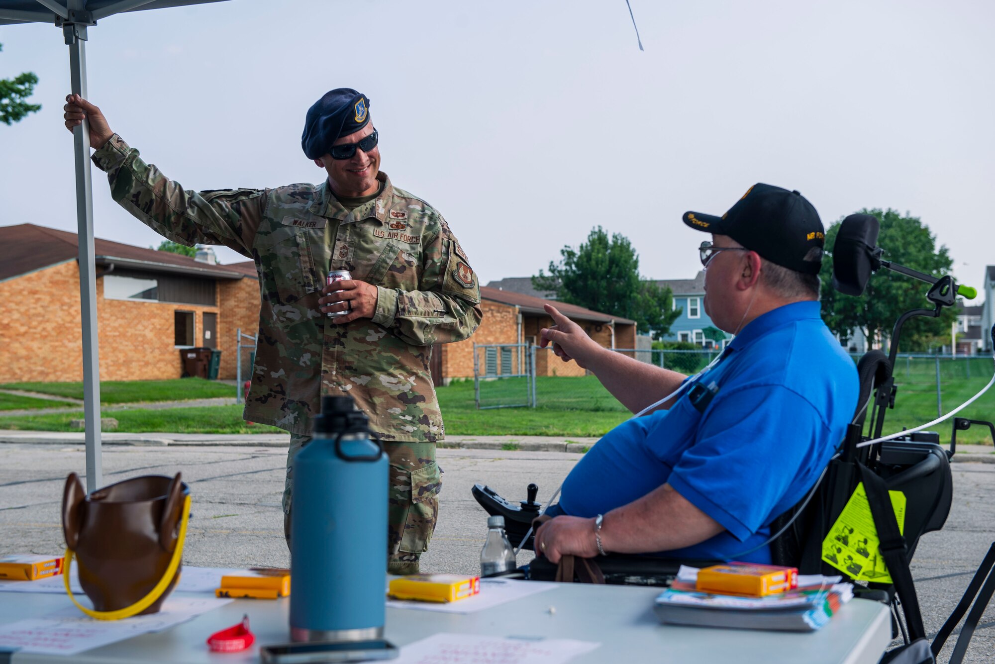 In front of a brown building with trees and grass surrounding it, a military member with a security forces black beret holds a pole in his right hand and a soda can in his right as he looks with a smile to a man in a wheelchair with a bright blue shirt and a black hat as they have a conversation under a tent.