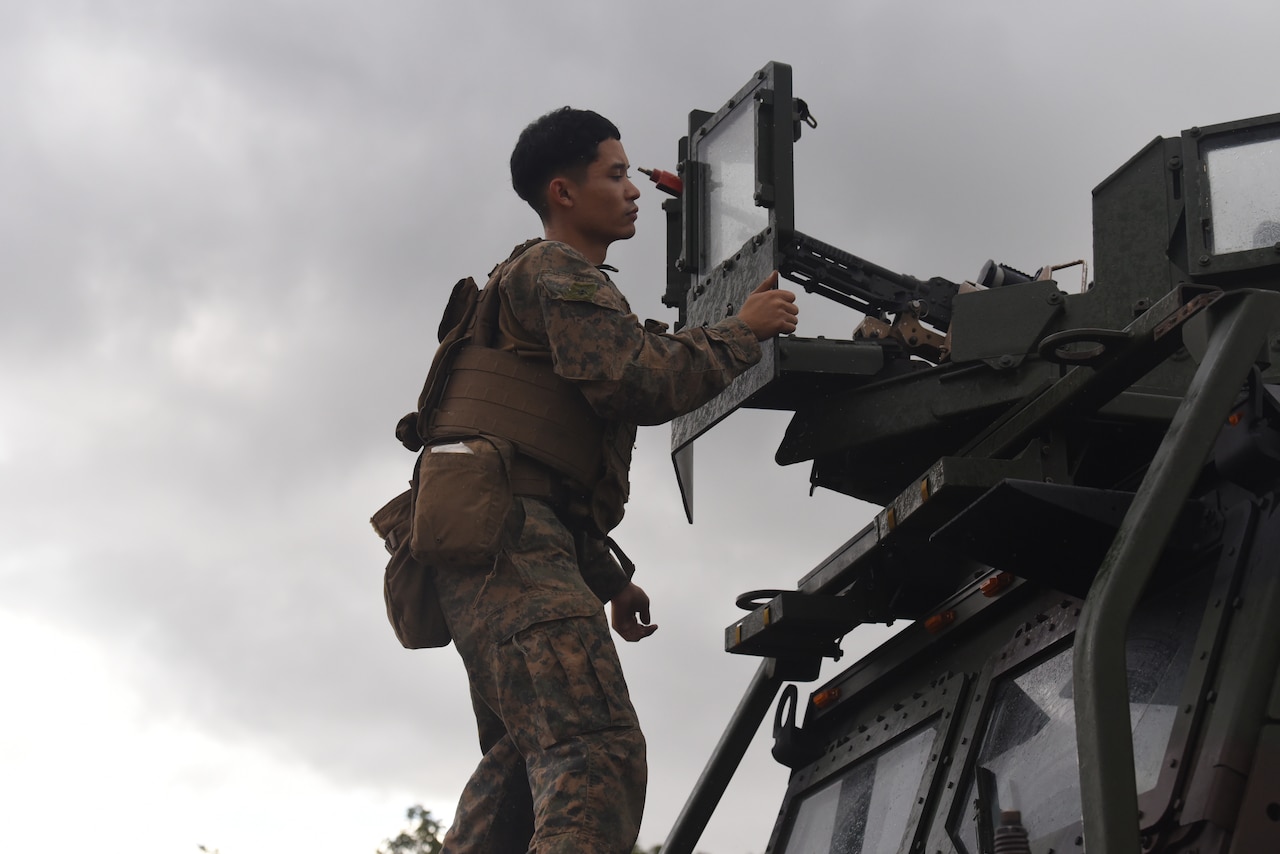 A uniformed service member stands on the hood of a military vehicle to adjust machinery attached to the roof.