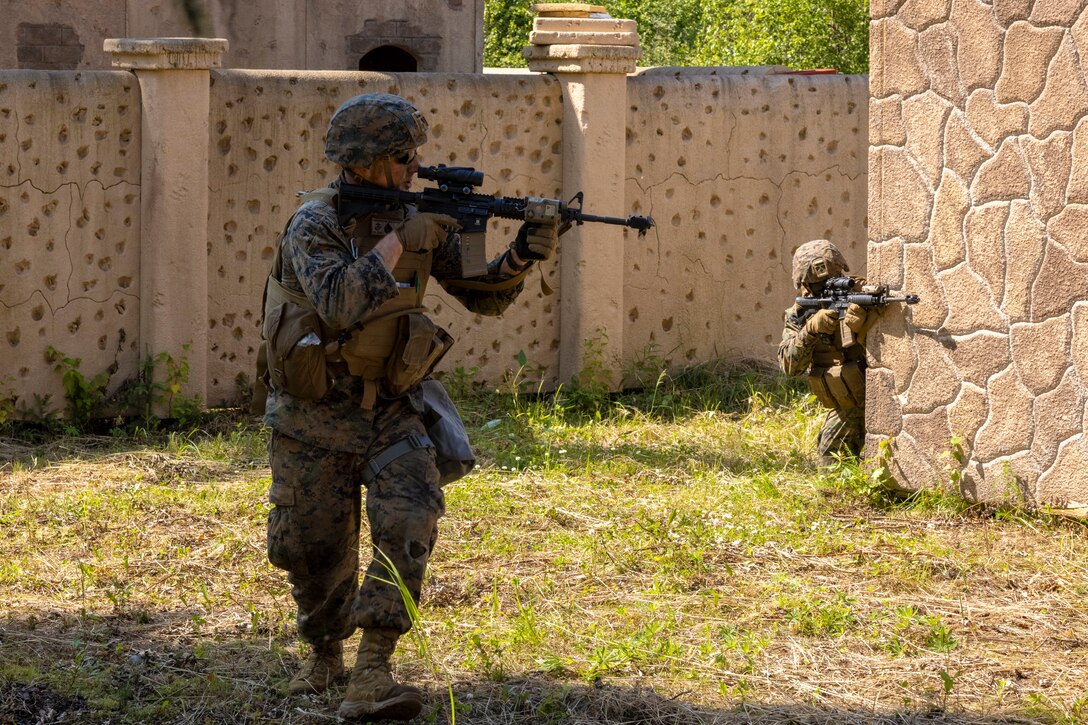 A Marine with a weapon kneels next a stone wall as a fellow Marine with a weapon walks on grass.