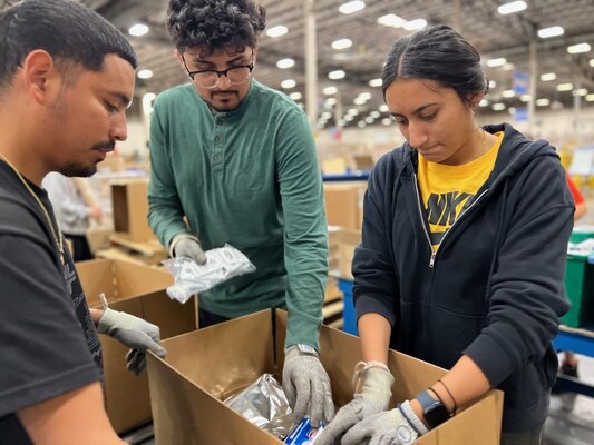 A group of interns put food items inside a e-rations container on an assembly line in a warehouse.