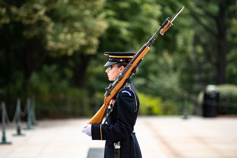 A tomb guard holds a weapon while walking along a pavement with trees in the backdrop.