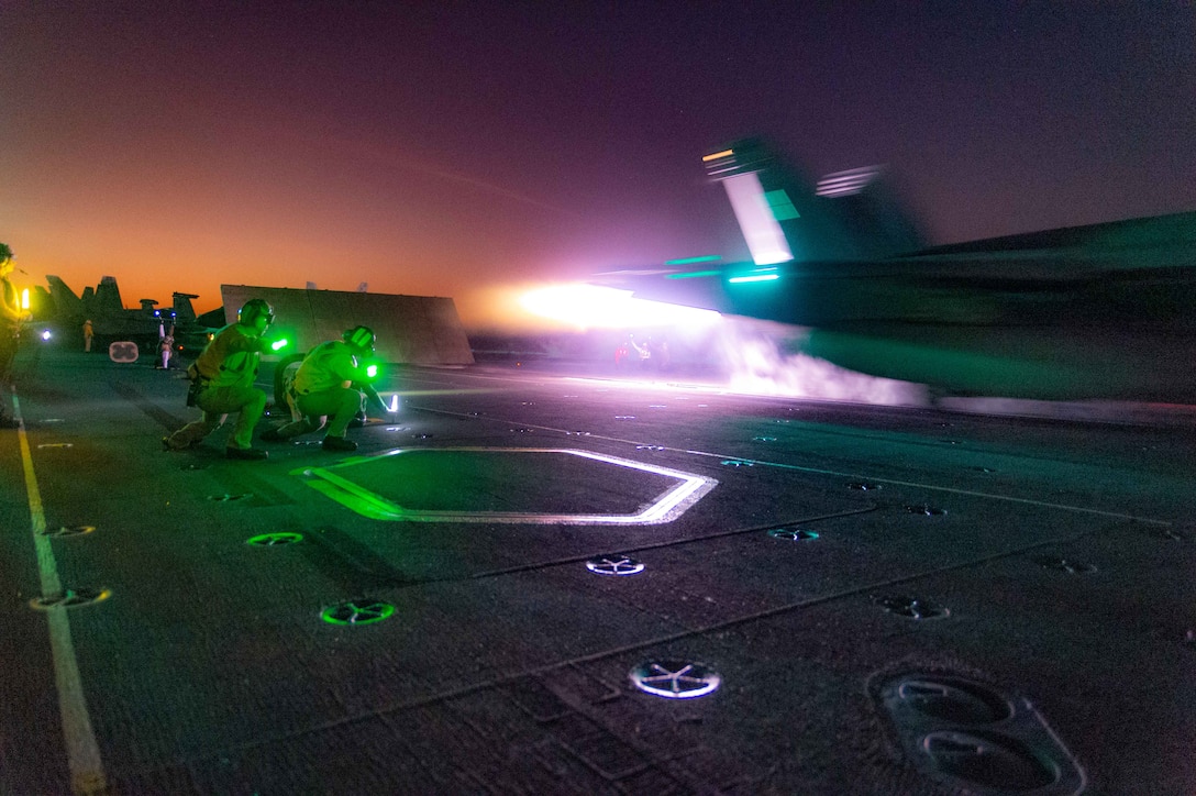 Two sailors crouch on the dark while giving signals as a jet takes off from a ship's deck with colored lights.