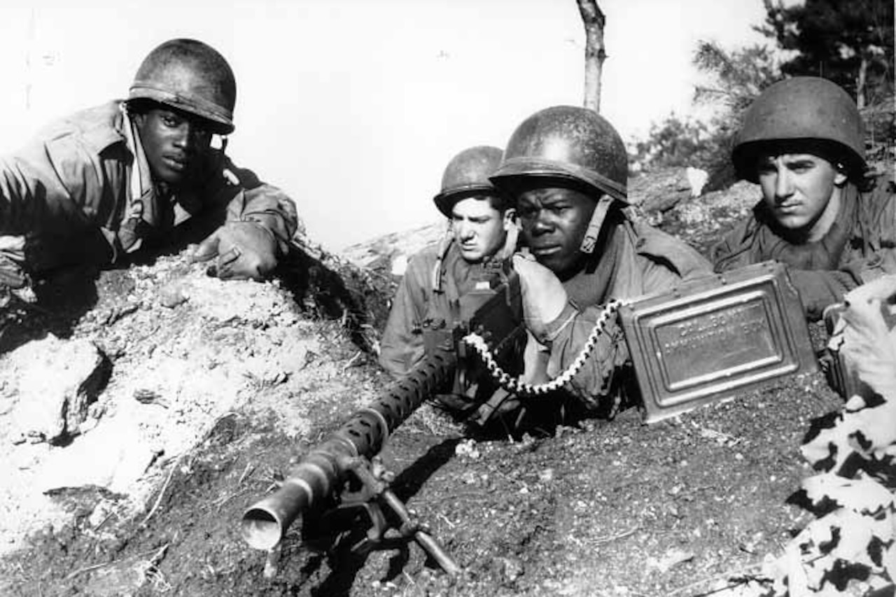 Four soldiers sit in a foxhole; one aims a weapon.