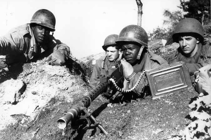 Four soldiers sit in a foxhole; one aims a weapon.
