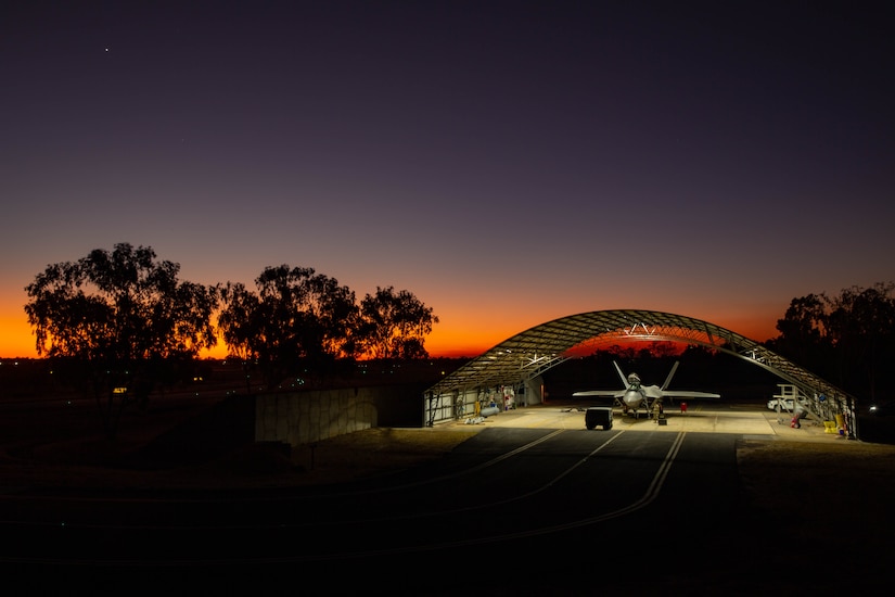 A fighter aircraft sits in a lighted hangar bay as the sun sets in the background.