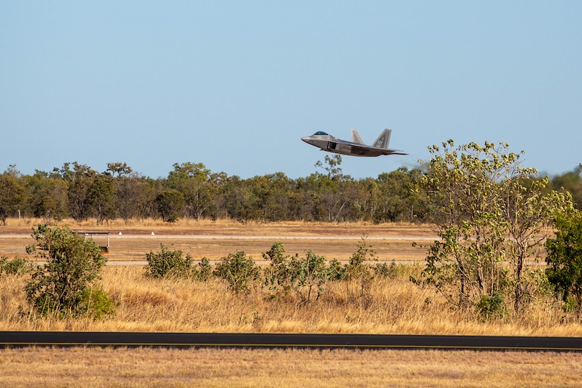 A fighter jet flies above a runway lined by trees.