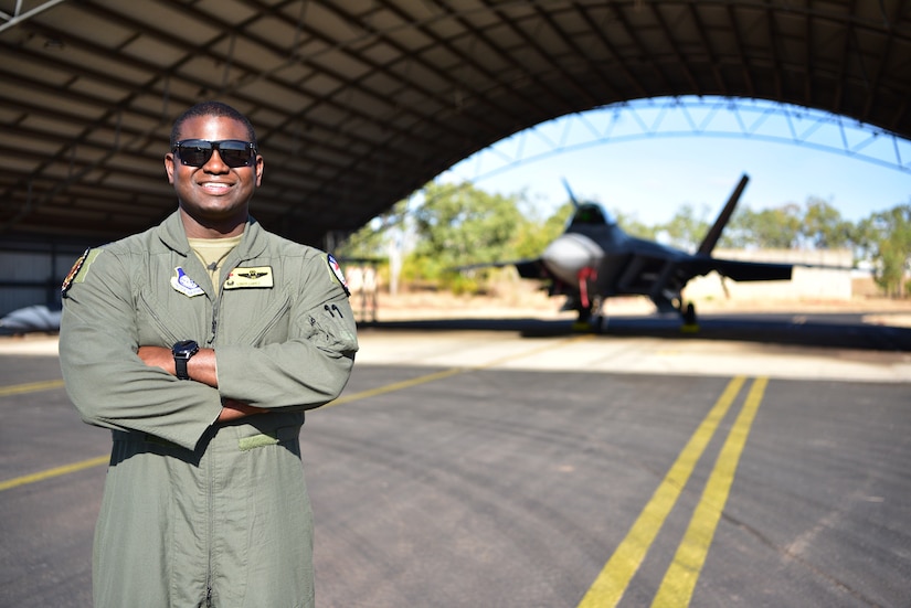 A man wearing a flight suit smiles as he stands with his arms crossed with a fighter jet in the background.