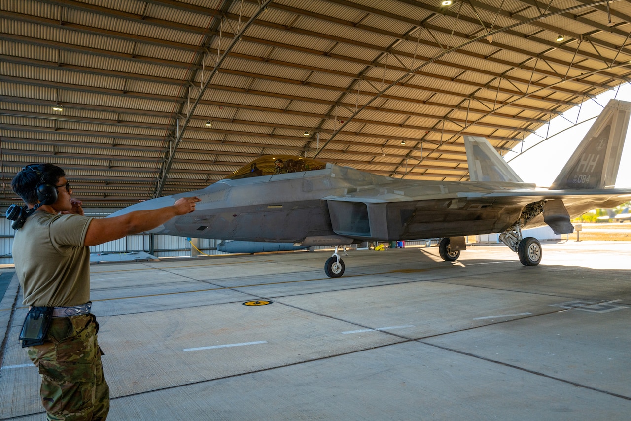 A service member in uniform points inside a hangar bay housing a fighter jet.