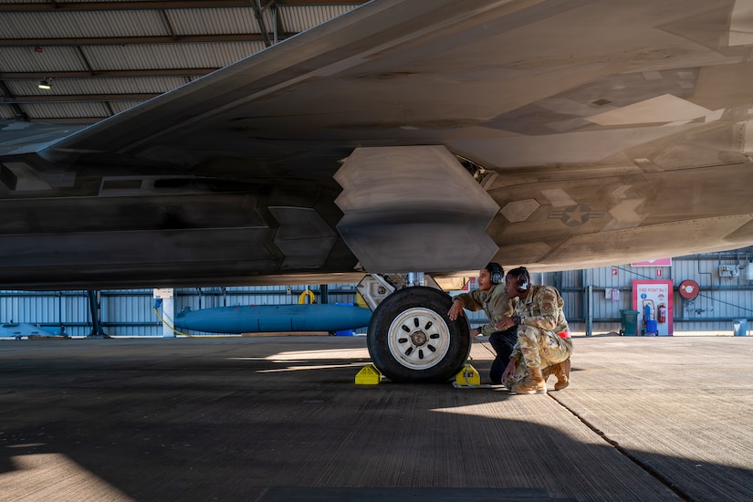 Two service members in uniform kneel beside the landing gear of a fighter jet.