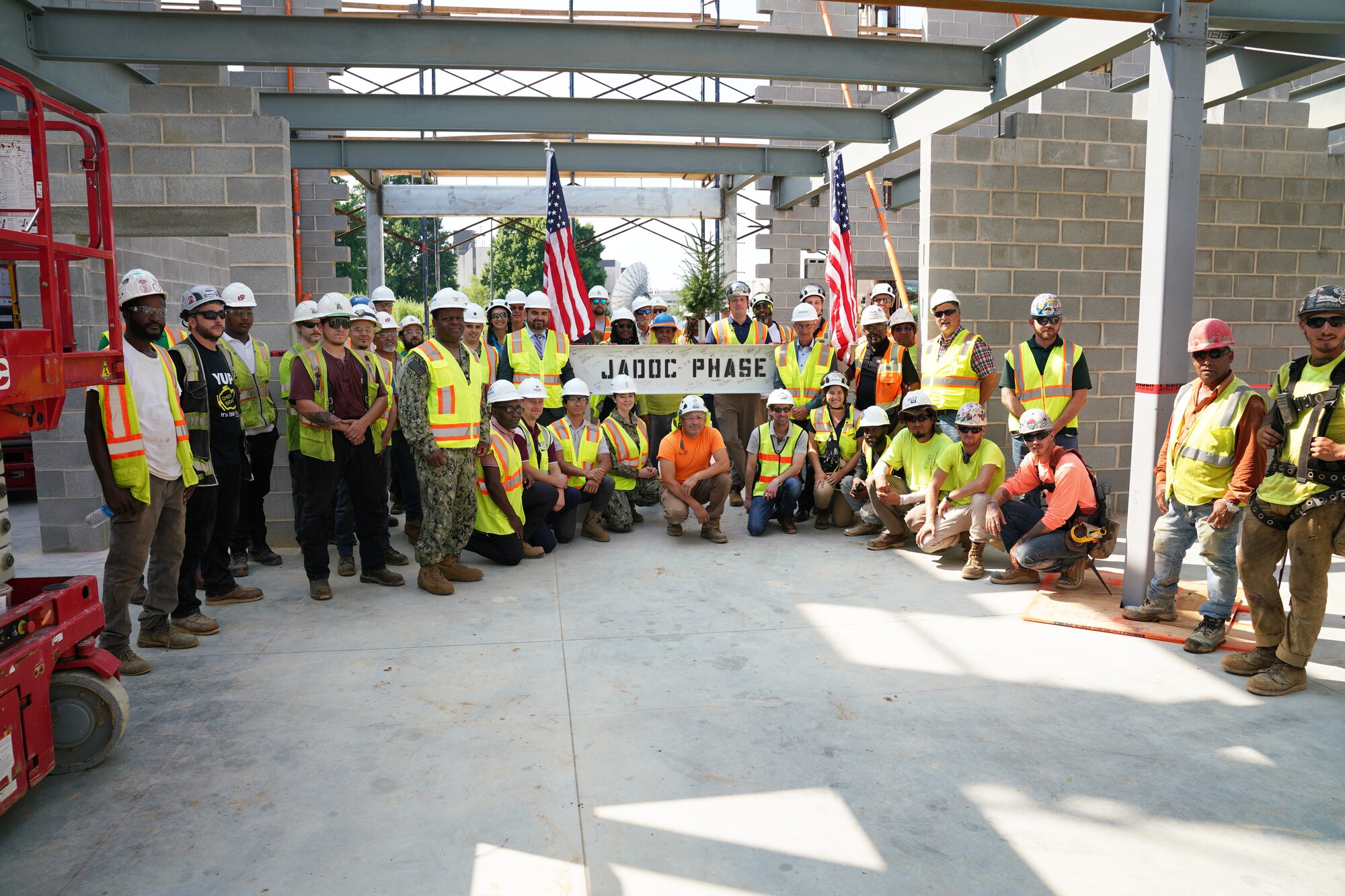 A group of construction workers and Navy officers pose in front of a steel beam on a construction site.