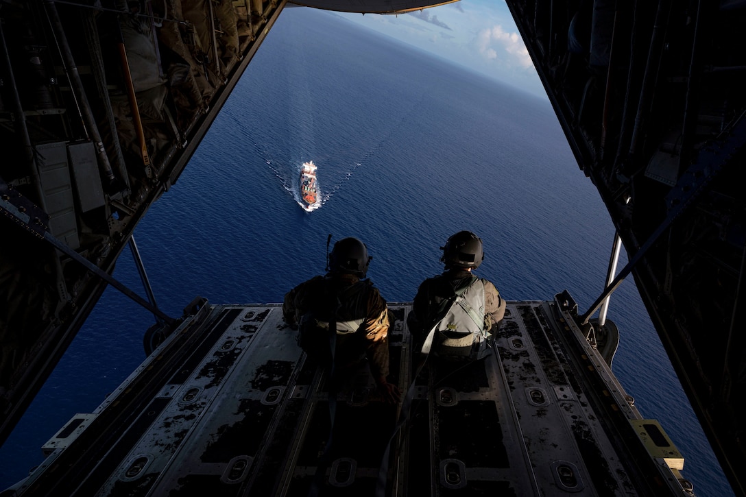 Two airmen watch a cargo ship transit a body of water while sitting next to each other in the open doorway of an airborne aircraft.