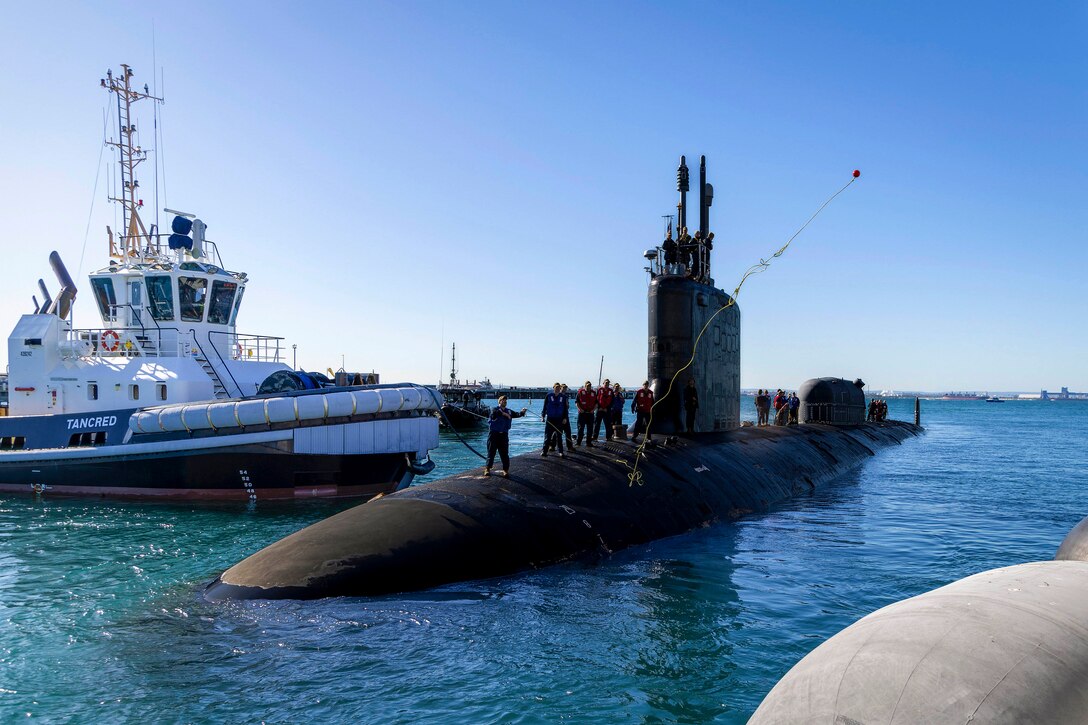A sailor heaves a line ashore from a submarine in a dock with a small boat on the other side.