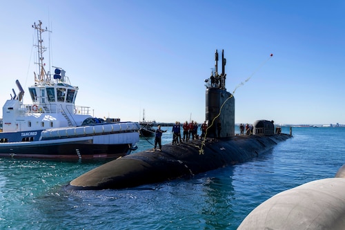 A sailor heaves a line ashore from a submarine in a dock with a small boat on the other side.