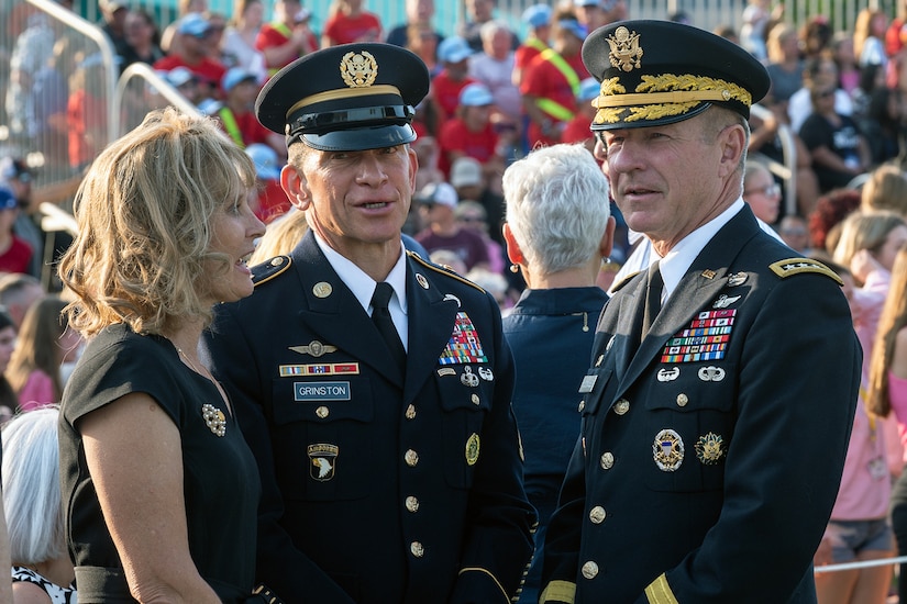 Two men and a woman stand and talk; a large crowd is in the background.