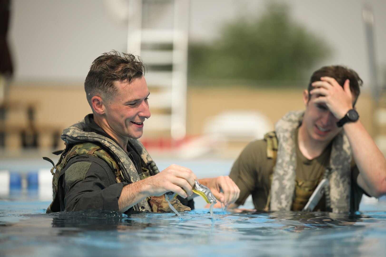 Two men in military uniform stand in chest-deep water during water survival training at a pool.