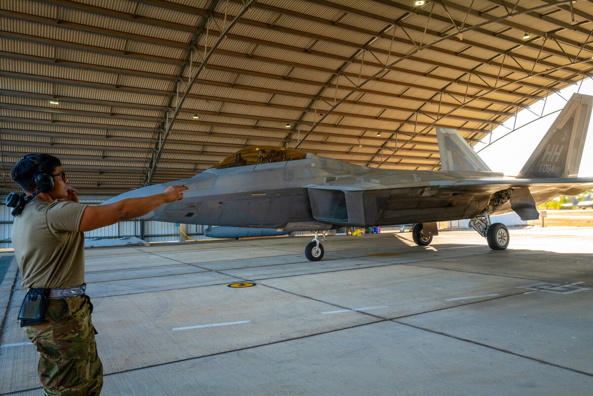 A service member in uniform points inside a hangar bay housing a fighter jet.