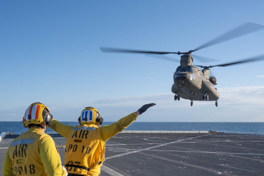One of two sailors in yellow puts out both arms while signaling for a  chinook helicopter to land on a ship's deck.