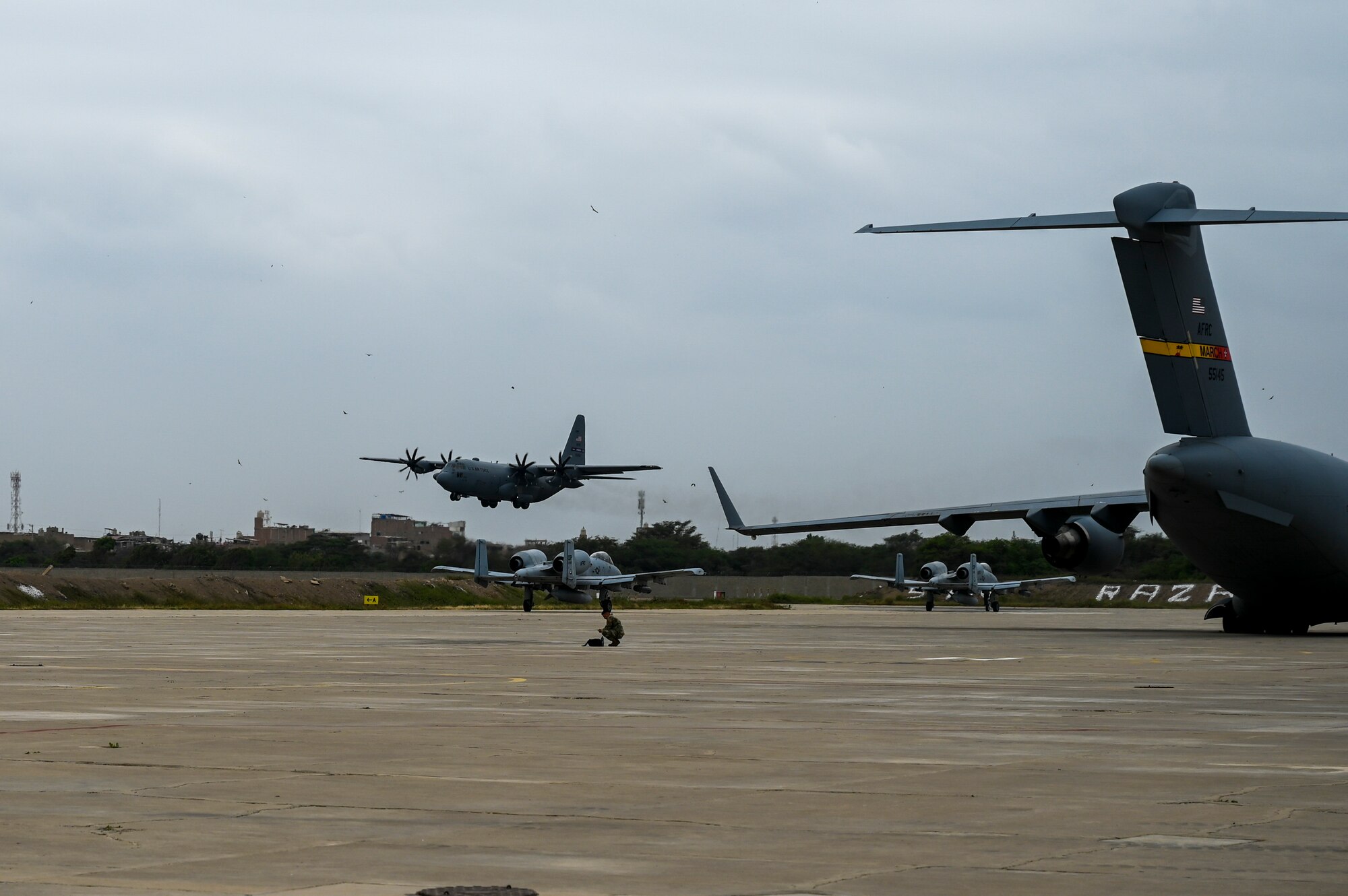 A C-130 lands on a runway behind two A-10s waiting to taxi out and a C-17 sitting on the flight line.