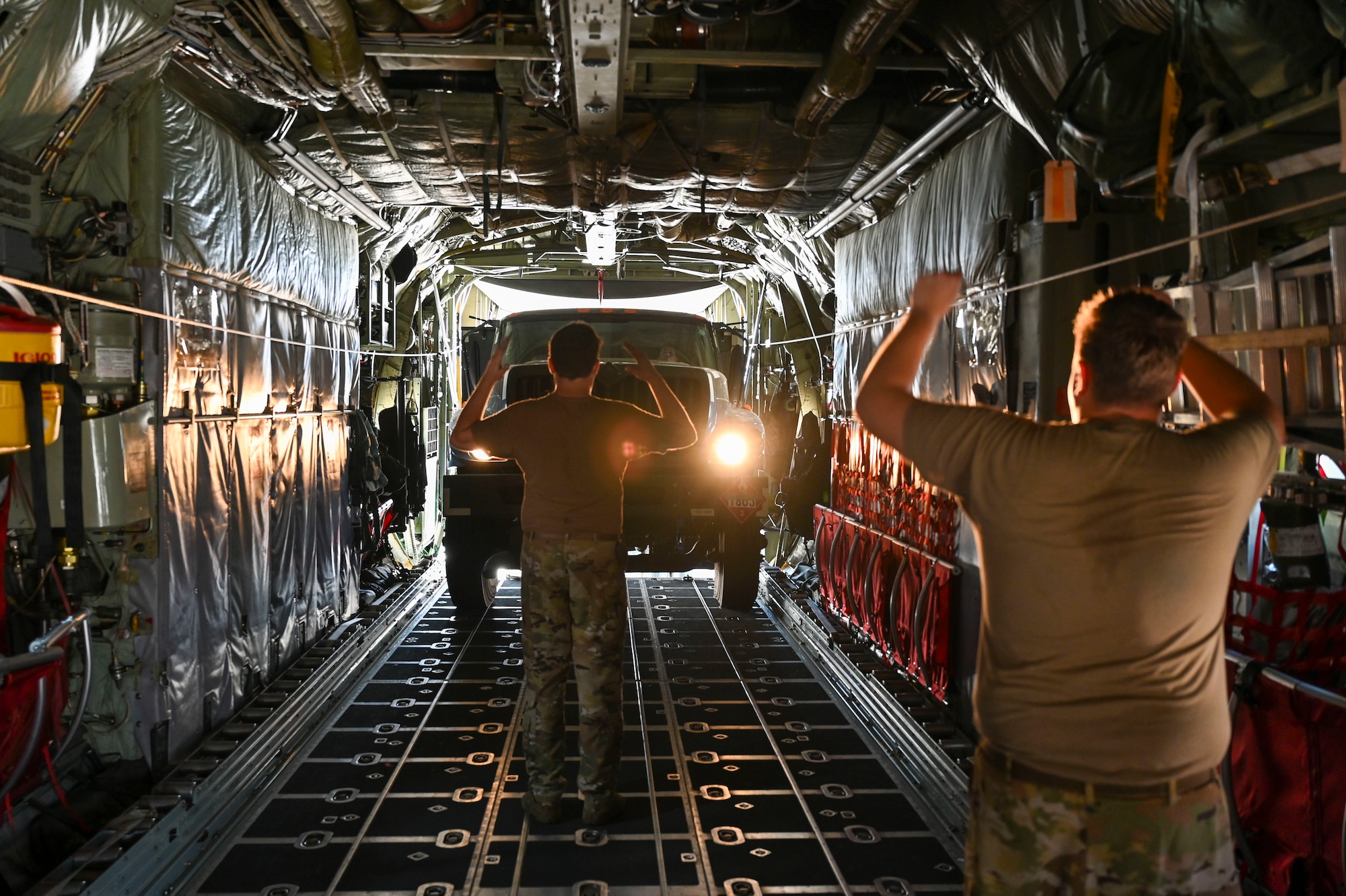 Two men use hand motions to marshal a fuel tanker truck up the ramp into the back of a C-130 cargo aircraft.