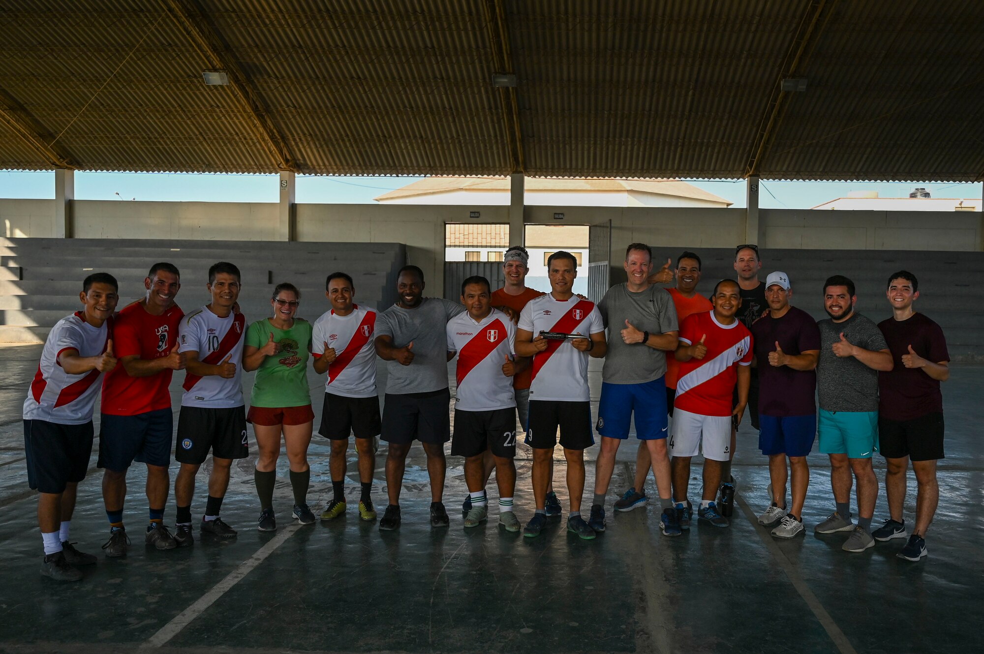 Five Peruvian Airmen wearing team jerseys line up intermingled with 9 American Airmen in mismatched shorts and t-shirts. All are giving the camera a thumbs-up sign.