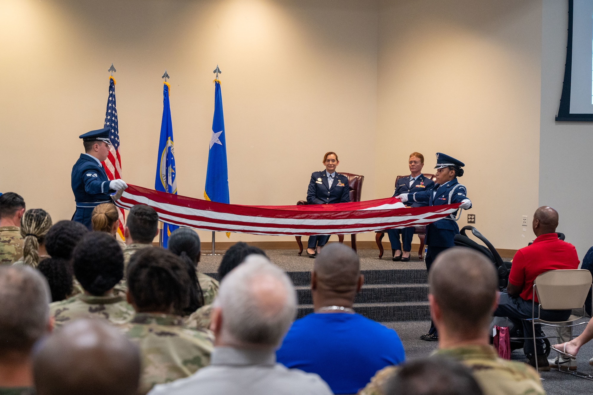 Two Airmen in service dress spread a U.S. flag out. Baldelli and Maj Gen Gunter sit behind watching. Crowd in the foreground also watches.