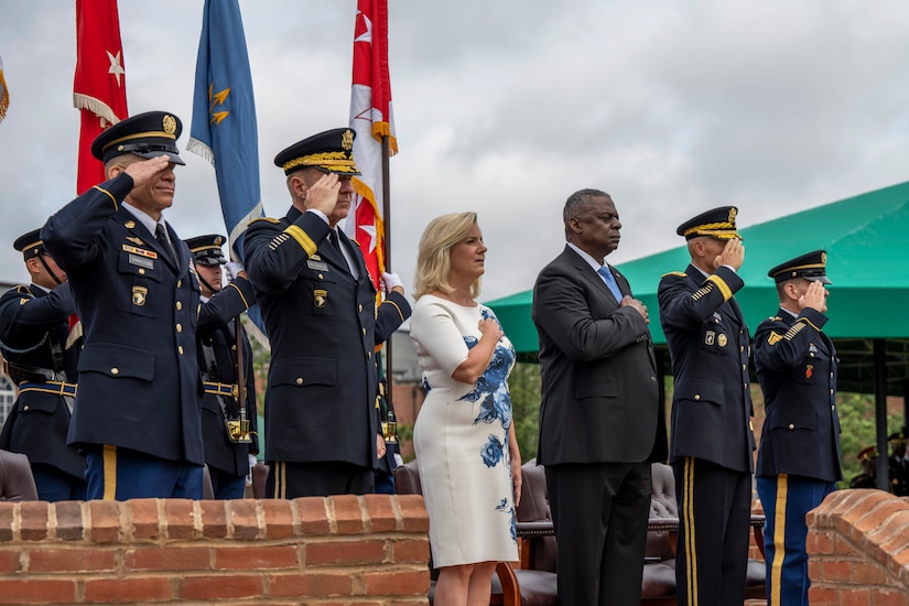 Military leaders stand in formation; some salute and others hold their right hand over their chest.