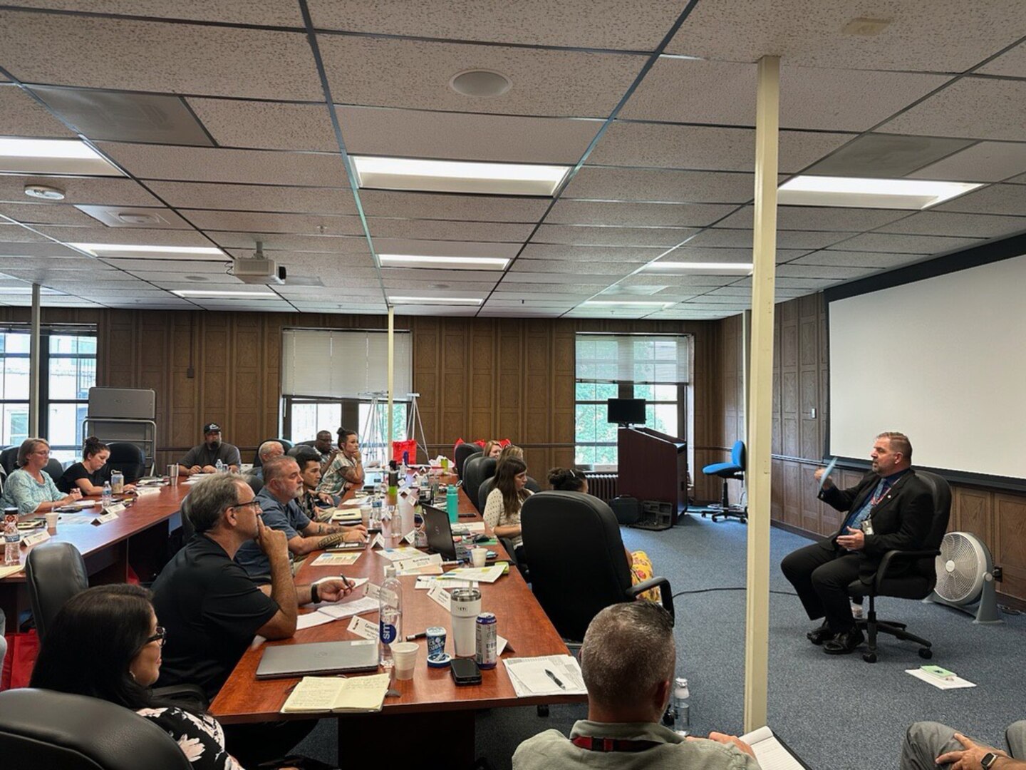 Man sitting in a chair in the front of the room while other people sit at tables.