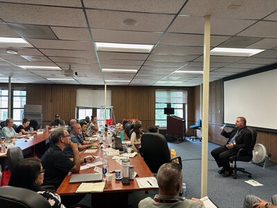 Man sitting in a chair in the front of the room while other people sit at tables.