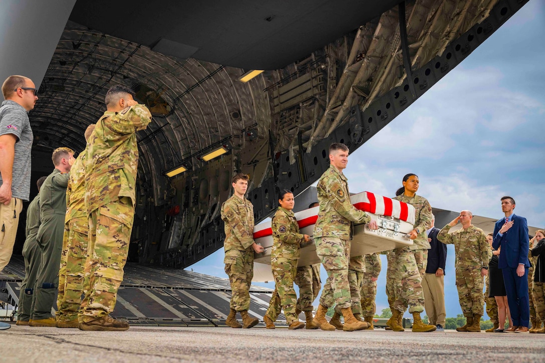 Airmen carry a casket draped in an American flag out of an aircraft as others stand in formation on either side.