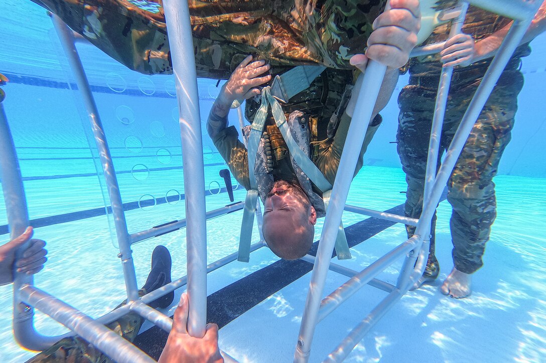 A uniformed airman holds their breath during underwater training while in a cage-like device.