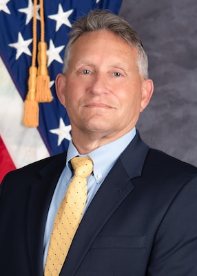 A gray-haired light skinned man (headshot) in a dark suit with a yellow tie and blue dress shirt.