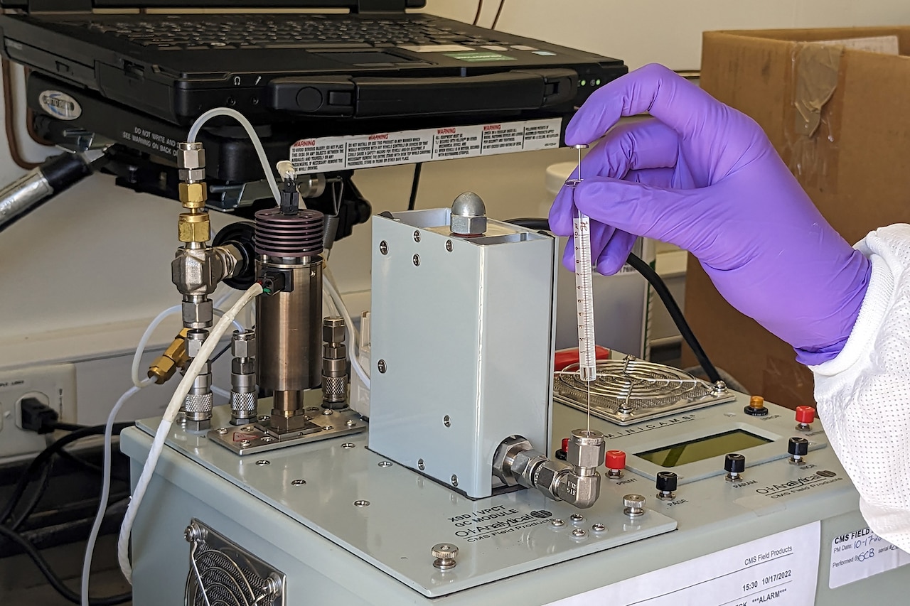 A hand plunges a syringe-like device into a gasket on a large piece of scientific equipment.