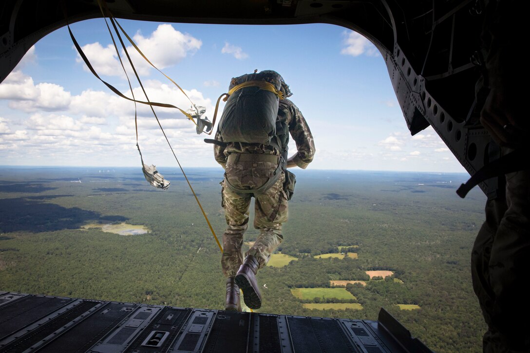 A British soldier wearing a parachute pack jumps out of the open doorway of an airborne aircraft with clouds and greenery in the distance.