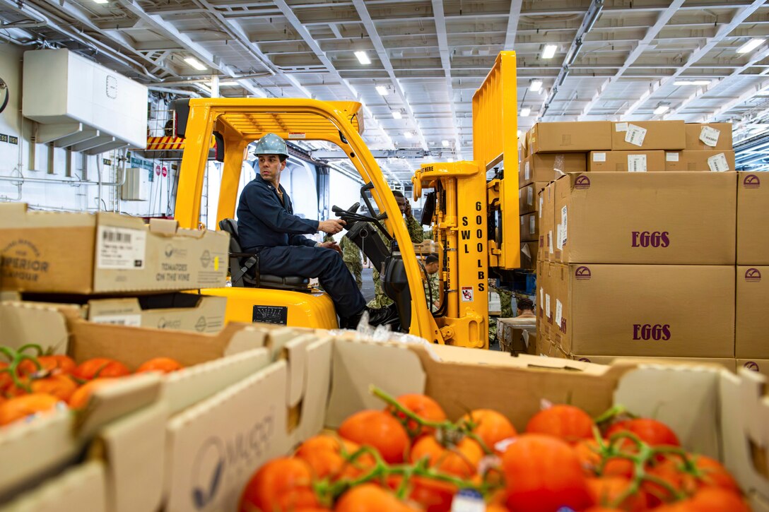 A sailor wearing protective gear operates a machine to move stacks of boxes as open boxes of tomatoes sit in the foreground.