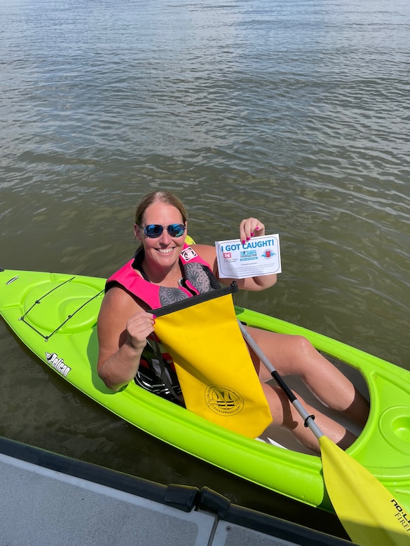 A woman in a pink life vest sits in a green kayak and hold up a piece of paper with water in the background.