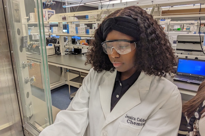 A scientist in a lab coat and protective glasses looks down at her work.