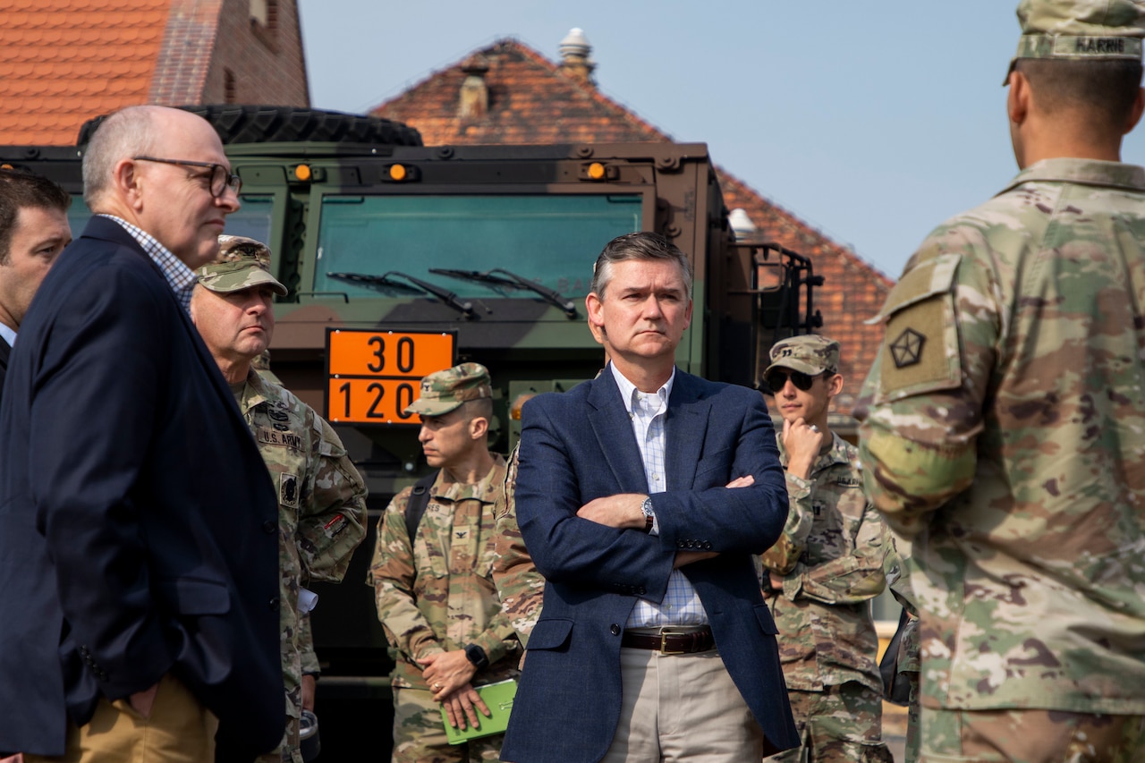 A man in a suit stands with his arms crossed as he speaks with military members in uniform.