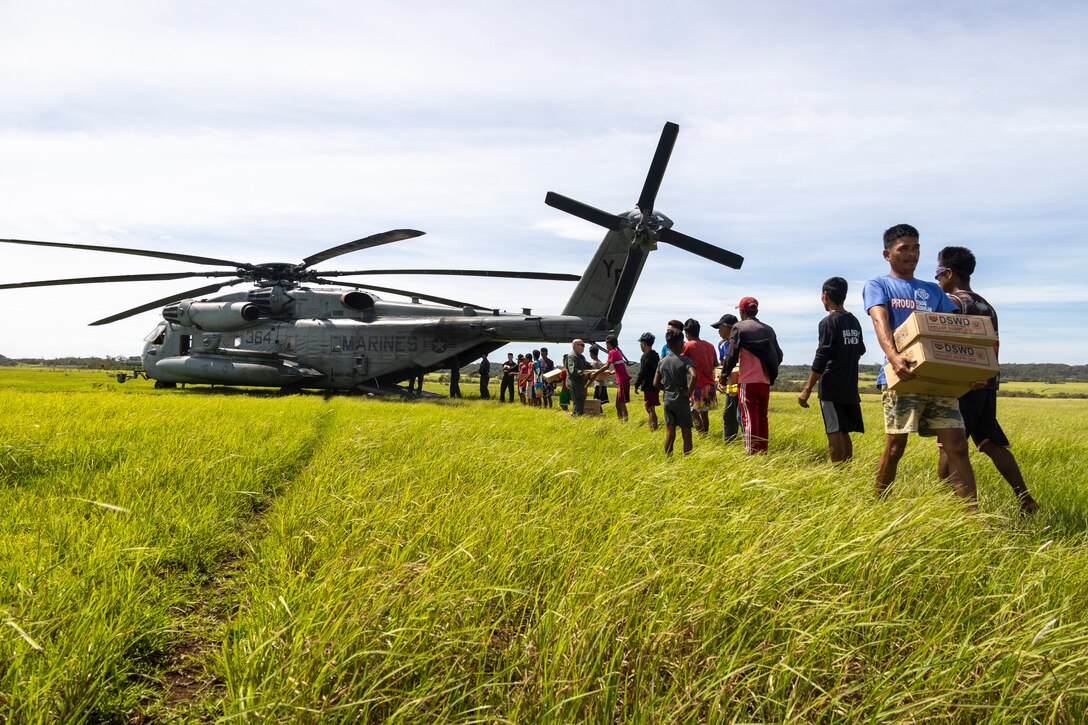 Service members and civilians carry boxes of supplies from a helicopter.