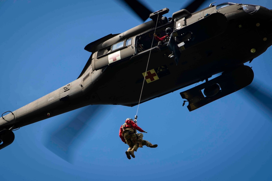 Service members are hoisted up by a rope attached to an airborne helicopter.