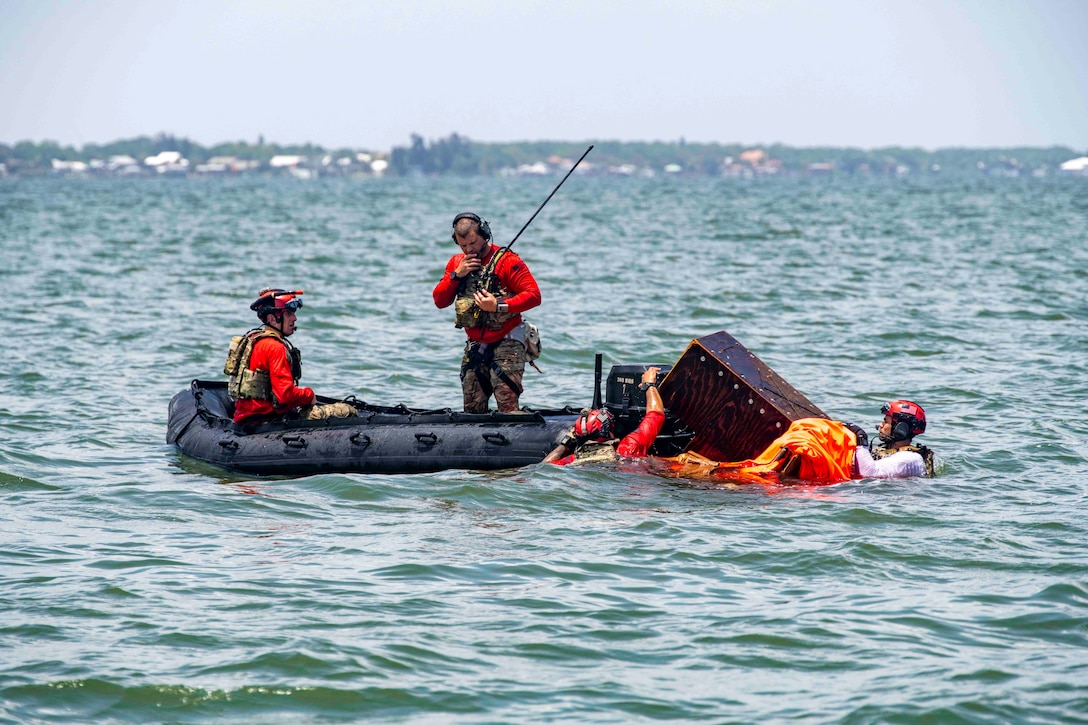 Two airmen stand and sit inside a small inflatable boat as others move an object in a body of water.
