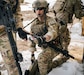 Service member kneels in the snow and reaches for small medical device that is a loaded autoinjector