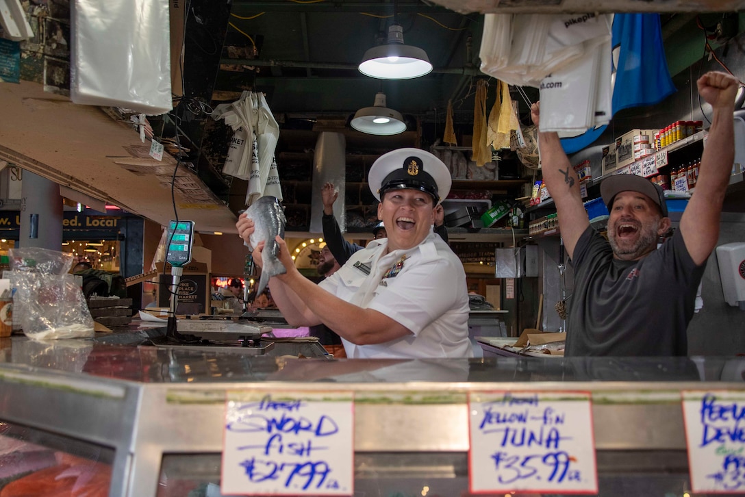 A smiling sailor catches a fish at a market as another person celebrates in the background.