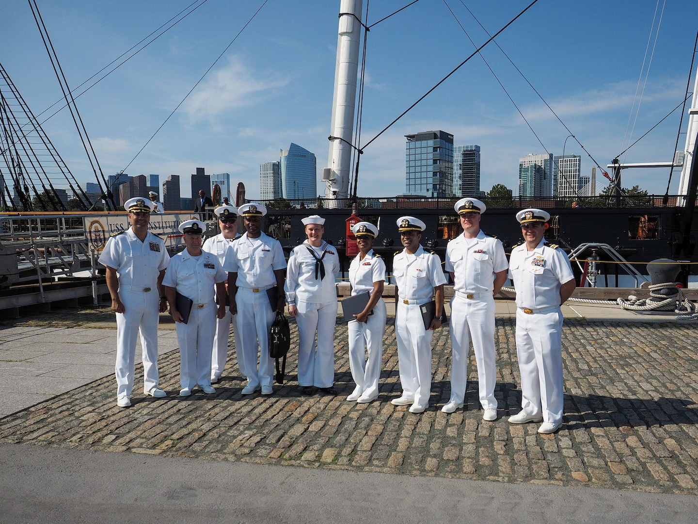 Nine sailors in white uniforms stand in front of a large sailing ship